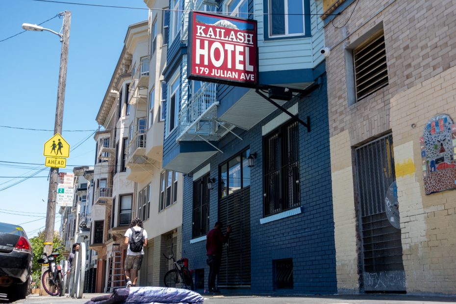 A person stands near the entrance of Kailash Hotel at 179 Julian Ave while another walks by on the sidewalk on a sunny day.