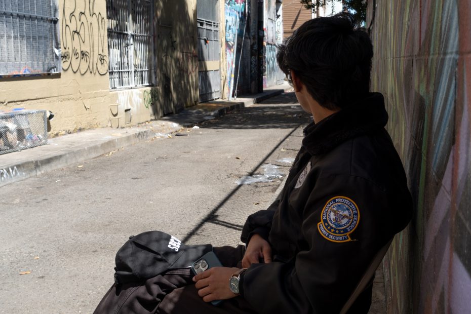 A person wearing a security jacket sits on a chair in an alleyway with graffiti-covered walls, looking away from the camera.