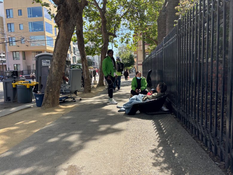 Three people are gathered on a sidewalk next to a black metal fence, with two sitting and one standing under a tree. Urban buildings and trash bins are visible in the background.