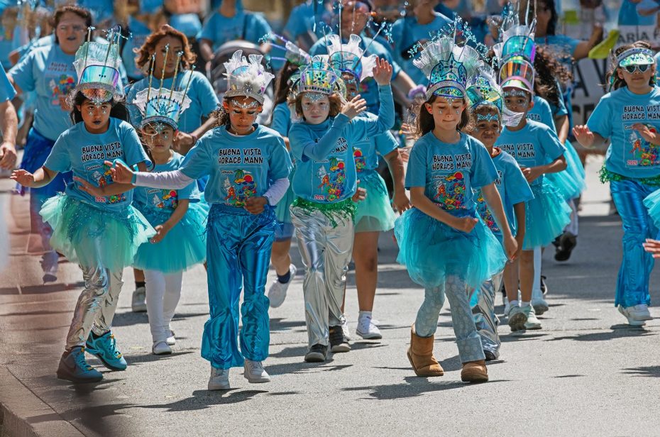 A group of children in colorful costumes and blue shirts parade down a street, wearing festive hats and tutus, participating in an organized outdoor event.