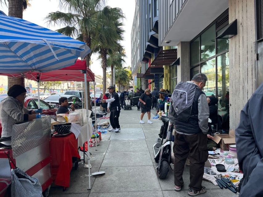 Street vendors and shoppers gather on a busy sidewalk lined with tables, food stands, and assorted goods in an urban setting.