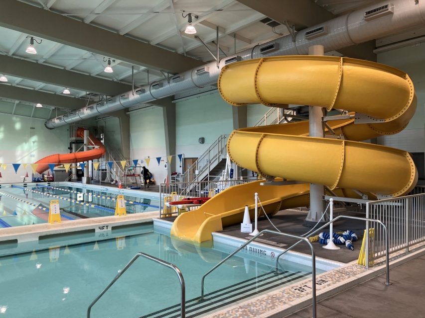 Indoor swimming pool area with lap lanes, a yellow spiral slide, an orange tube slide, and pool safety signs. The space is well-lit with a high ceiling and ventilation ducts.