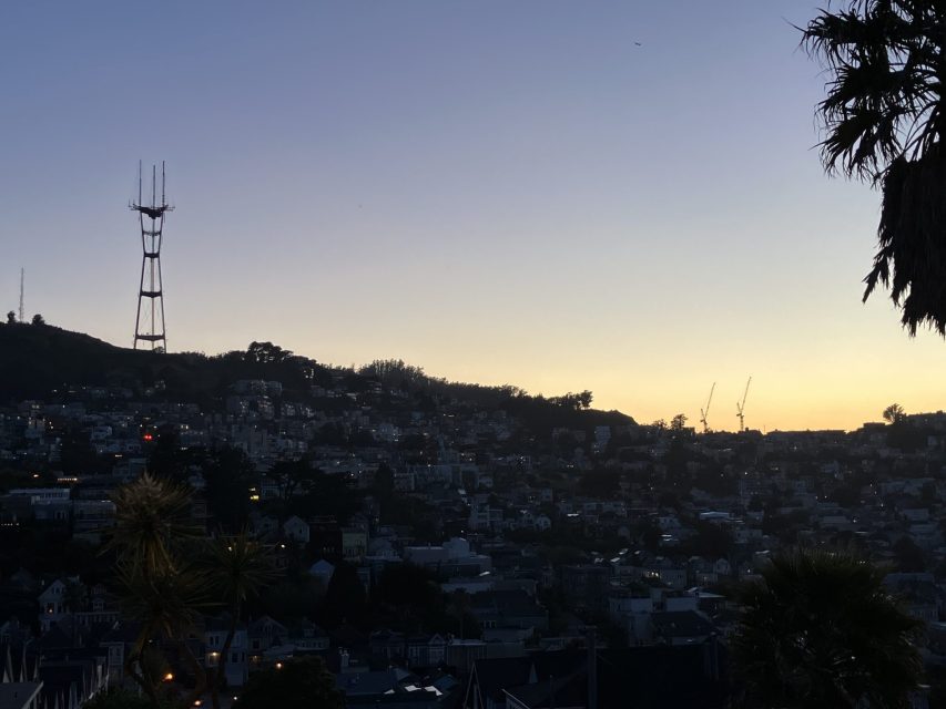 A cityscape at dusk features a hill with a tall radio tower, residential buildings, construction cranes, and silhouetted trees against a fading sky.