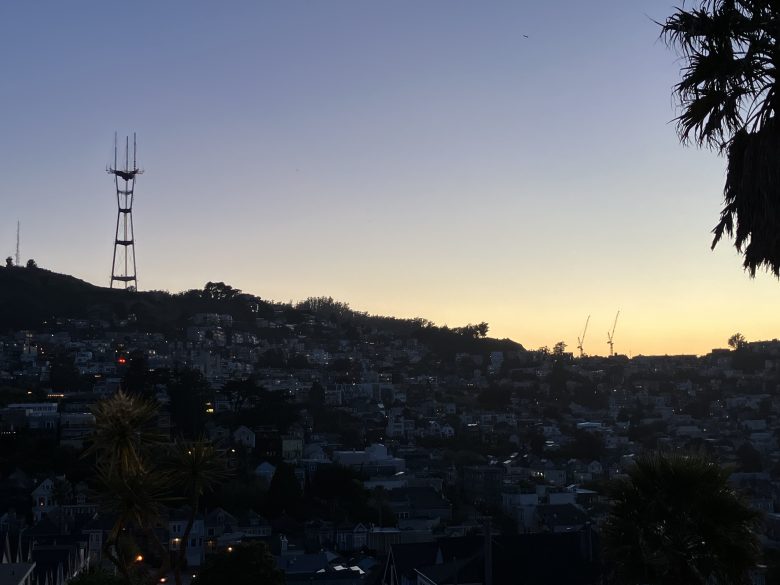 View of a city hillside at dusk with Sutro Tower on the left, construction cranes on the right, and palm trees silhouetted in the foreground.