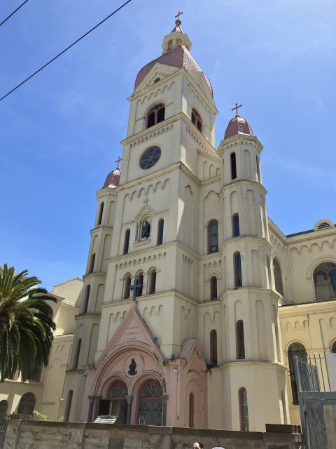 A cream-colored church with two tall towers, arched windows, and red domes, set against a clear blue sky with a palm tree on the left.