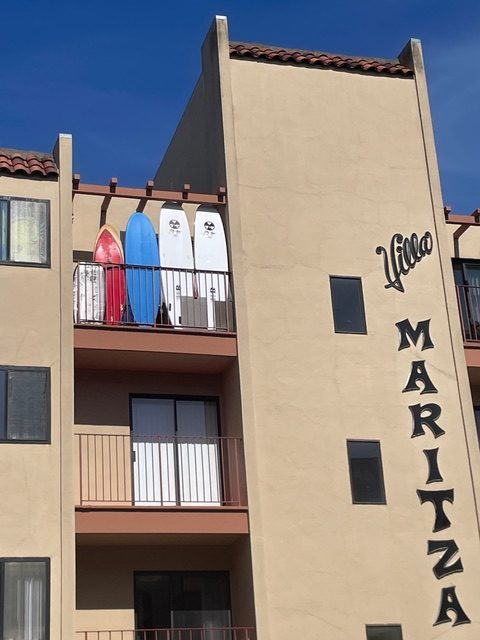 Four surfboards are standing upright on the balcony of an apartment building named "Villa Maritza" with beige walls and a red tile roof.