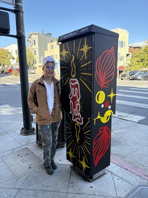 A person stands next to a painted utility box featuring a red candle, yellow rays, and red and yellow abstract designs on a city street corner.