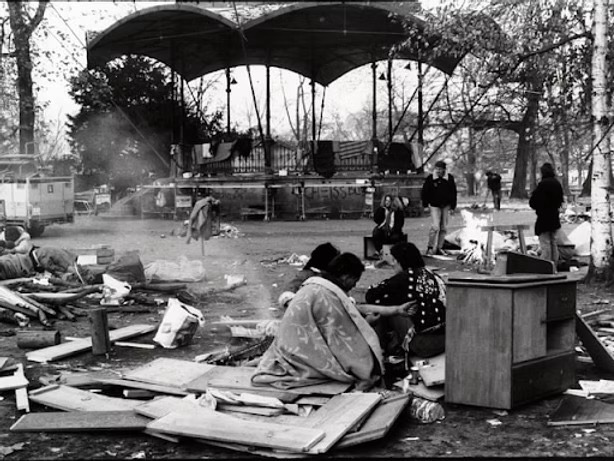 A group of people sits among scattered debris and furniture in an outdoor area near a stage structure, with trees and other individuals in the background.
