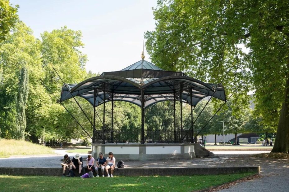 Five people sit on a low wall in front of an ornate gazebo surrounded by trees in a sunny park.