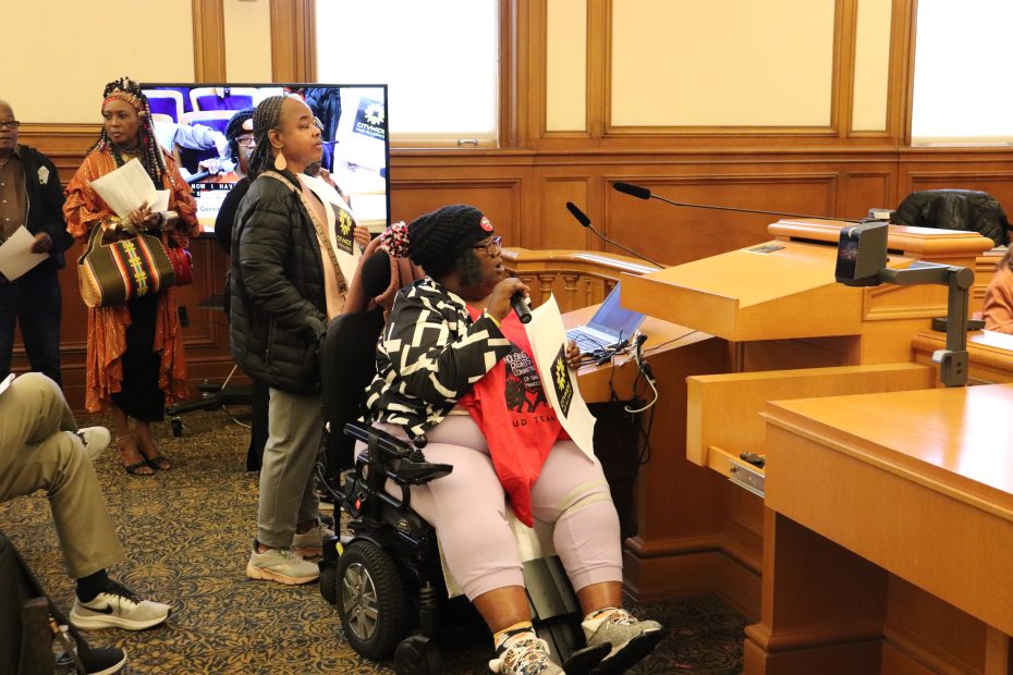 A woman in a wheelchair speaks at a podium in a wood-paneled room while others stand nearby, with a TV screen in the background displaying the scene.