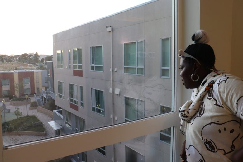 A person stands indoors looking out of a large window at a modern apartment building and courtyard during daylight.