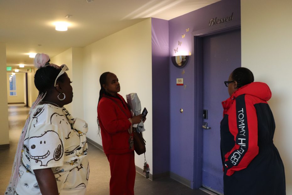 Three women stand and talk in the hallway of an apartment building in front of a purple door labeled "Blessed" with gold butterfly decals.