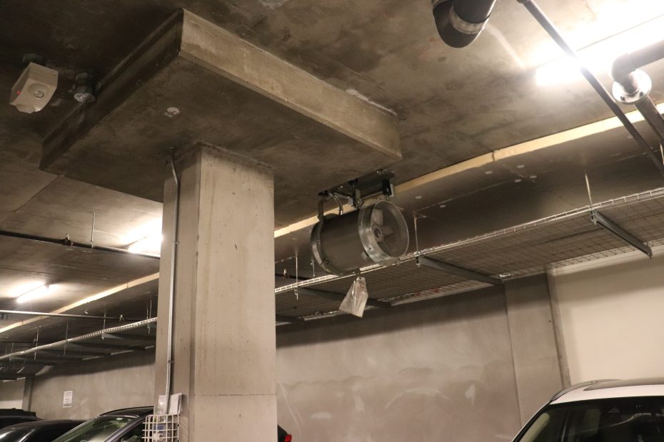 A metal keg is suspended from the ceiling by chains in an underground parking garage, near concrete support columns and parked cars.