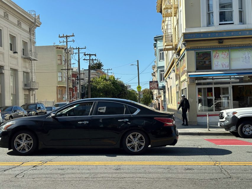A black sedan is parked on a city street with buildings, a hotel sign, and a person walking on the sidewalk in the background under clear blue skies.