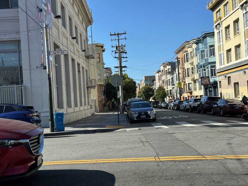 A city street with parked cars on both sides, buildings lining the street, a stop sign, and a clear blue sky overhead.