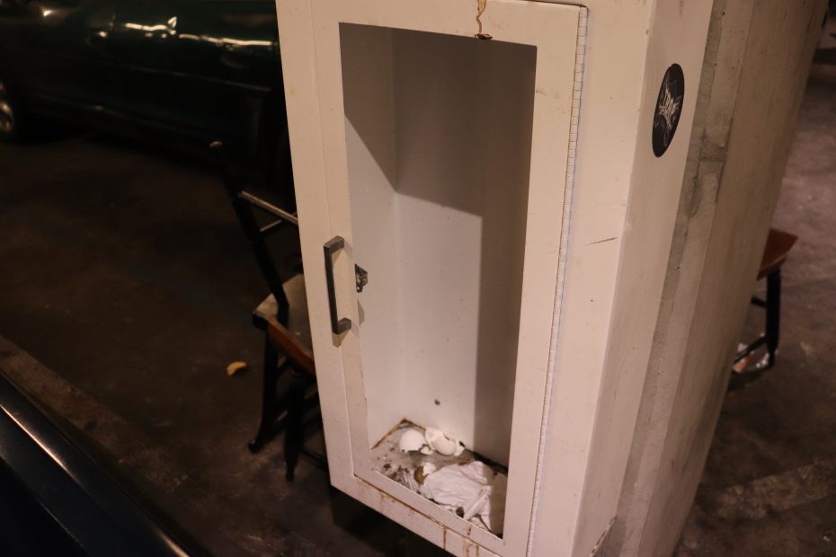 An empty wall-mounted fire hose cabinet with trash and debris inside, located in a parking garage.
