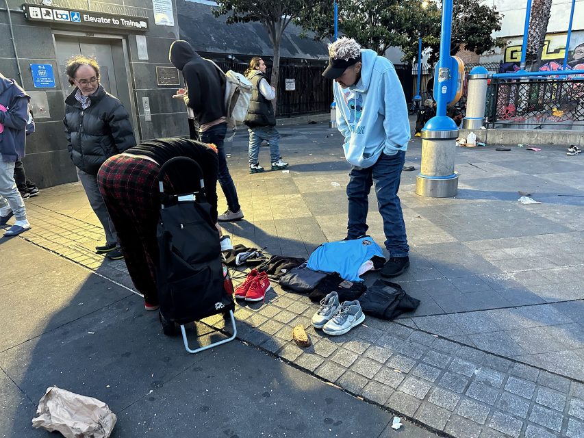 Several people gather around clothing and shoes spread on the sidewalk near a metro entrance in an urban area.