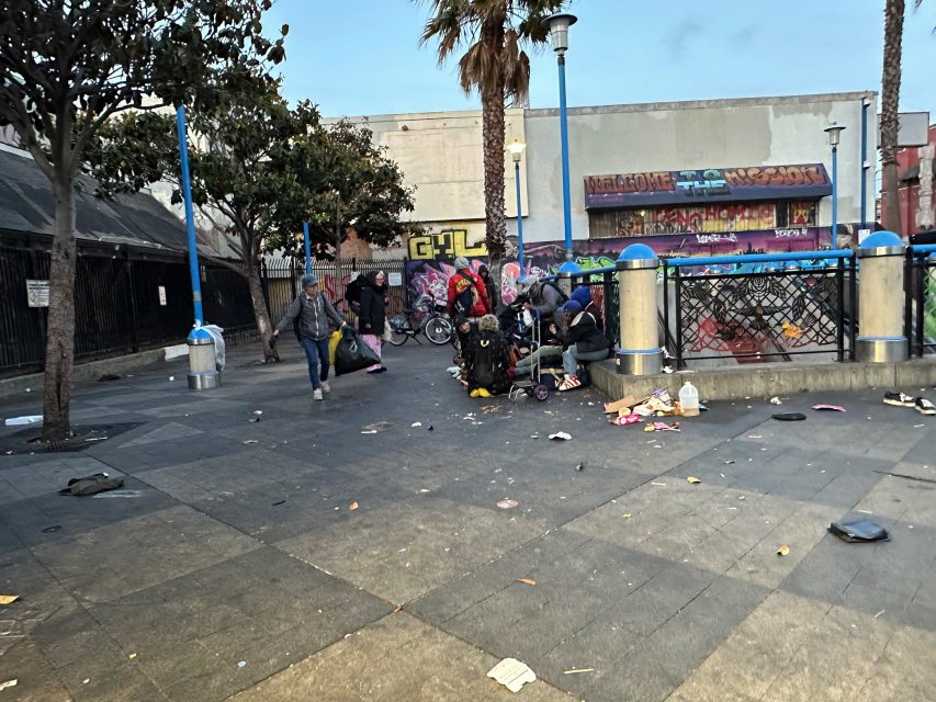 A group of people gather beside a fence in an urban plaza littered with trash; graffiti covers a building in the background.