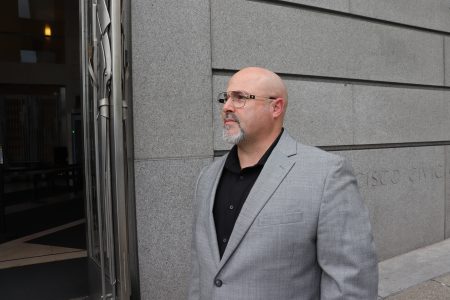 A man in a gray suit and black shirt stands outside a building with a stone facade, looking to the side.