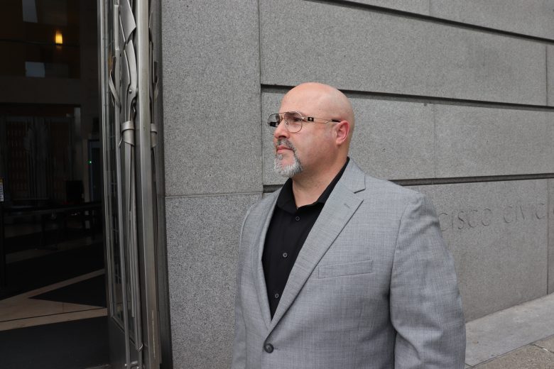 A man in a gray suit and black shirt stands outside a building with a stone facade, looking to the side.
