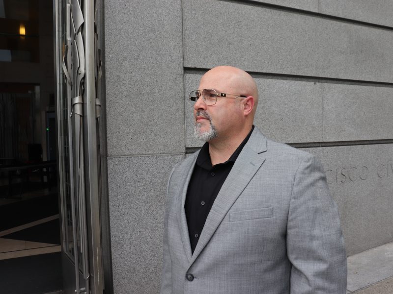 A man in a gray suit and black shirt stands outside a building with a stone facade, looking to the side.
