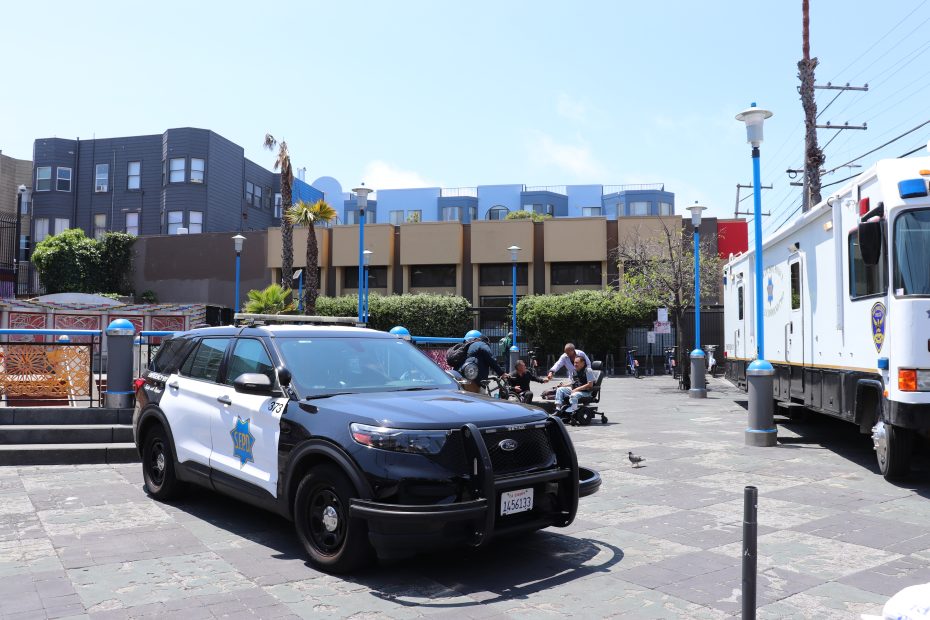 A parked police SUV and a large police vehicle are in a city plaza. Two people sit on benches in the background, and buildings line the street under a clear sky.