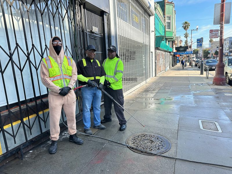 Three workers in safety vests stand on a city sidewalk near a gate, holding cleaning tools. Urban buildings and pedestrians are visible in the background.