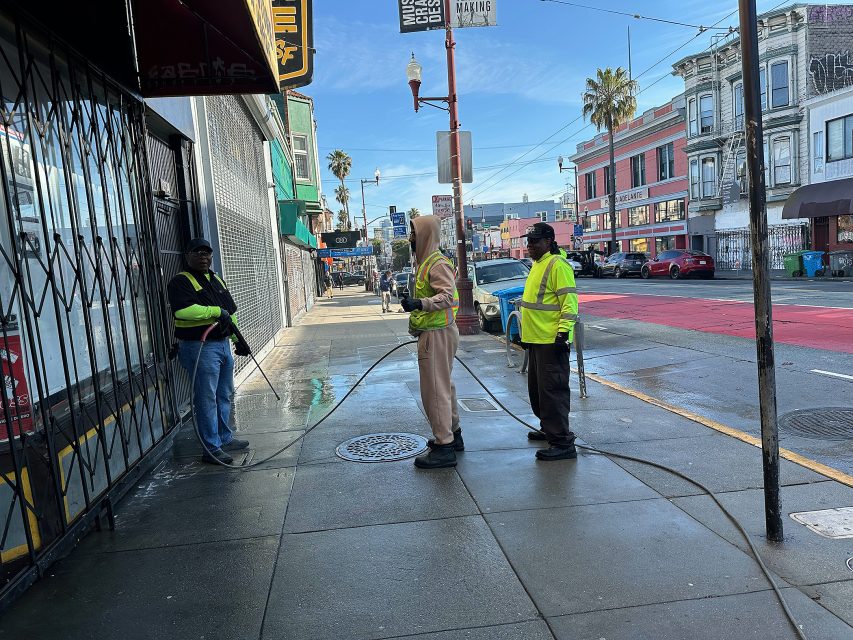 Three people in reflective safety vests stand on a city sidewalk, cleaning the pavement with a hose and pressure washer. Urban buildings and palm trees line the street in the background.