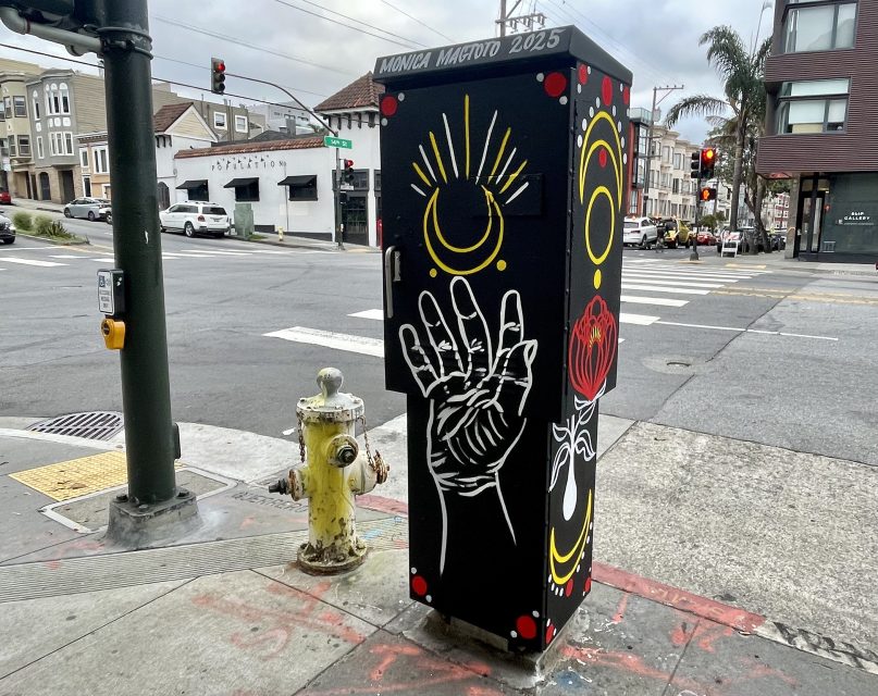 Traffic utility box at a city intersection painted with a hand, a yellow crescent moon, sun rays, a red rose, and the name "Monica Magtoto 2025" on the top edge.