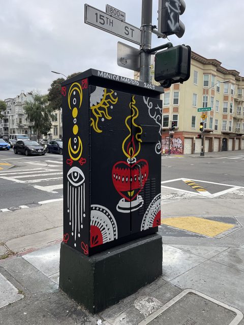 A painted utility box on a street corner features red, yellow, and white abstract designs, including an eye and patterns, with "Monica Magoto 2025" written at the top.