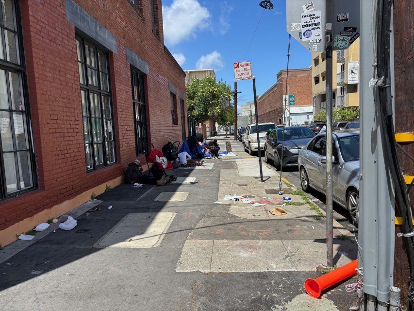 Several people sit or lie on a city sidewalk next to a brick building, surrounded by scattered belongings and litter, with parked cars lining the street.