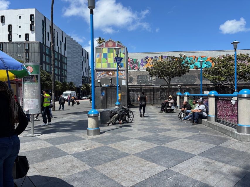 A city plaza with people sitting on benches, a worker in a safety vest, a person in a wheelchair, and buildings with graffiti in the background under a partly cloudy sky.