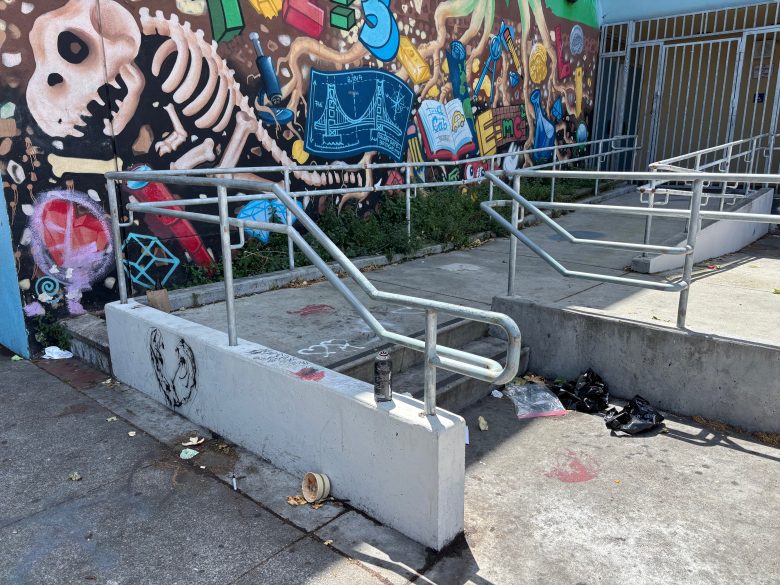 A concrete wheelchair ramp with metal railings and scattered trash in front of a colorful mural featuring skeletons and abstract designs.
