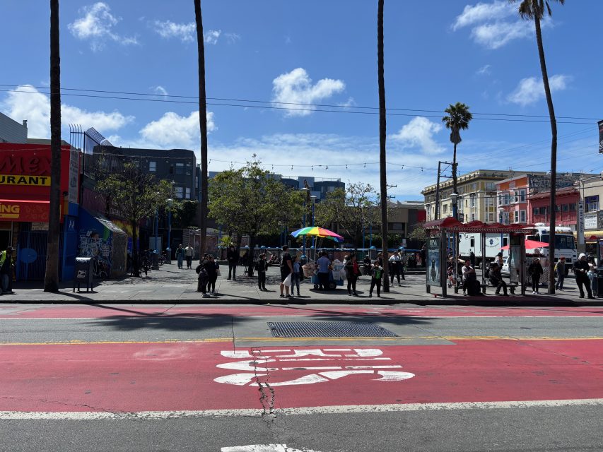 A city street with people gathered in a plaza, food trucks, a rainbow umbrella, palm trees, and colorful buildings under a blue sky with clouds.