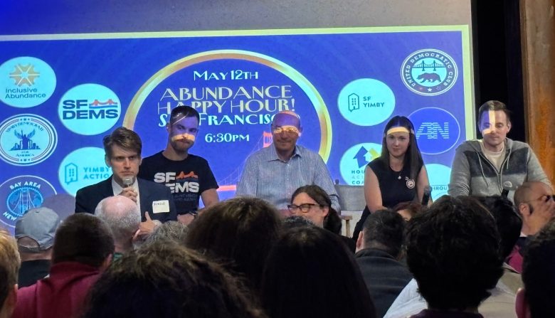 A panel of five people sits in front of a screen displaying event details and organization logos at an indoor gathering with an audience.