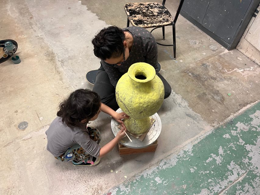 An adult and a child work together on a large yellow pottery vase in a workshop, with tools and a skateboard visible nearby.