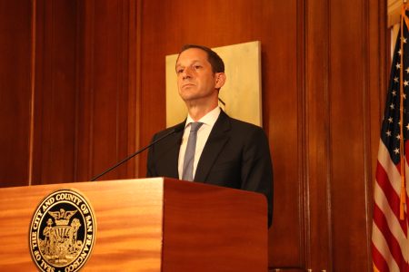 A man in a suit stands at a podium with an official seal, speaking in a wood-paneled room with a U.S. flag in the background.