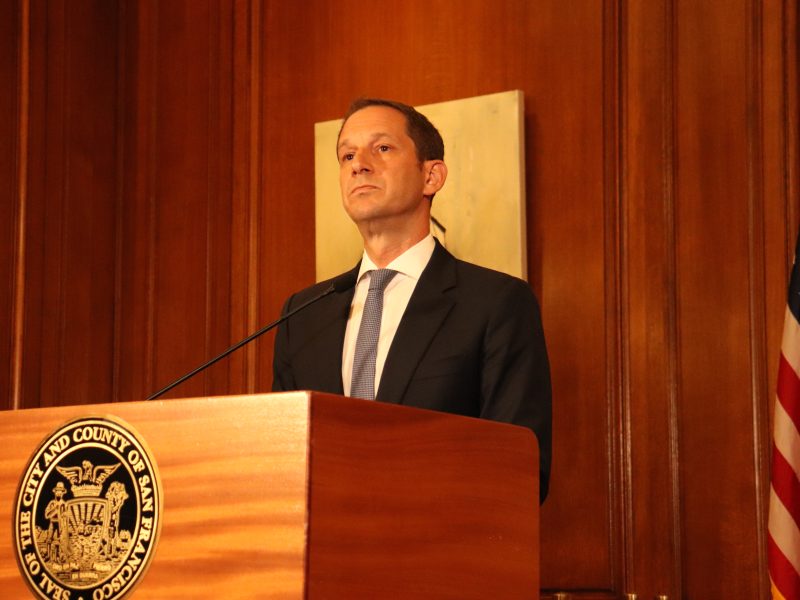 A man in a suit stands at a podium with an official seal, speaking in a wood-paneled room with a U.S. flag in the background.