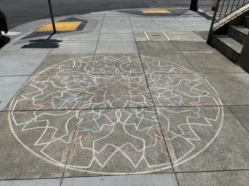 A large, intricate mandala design is drawn with colored chalk on a concrete sidewalk at a street corner.