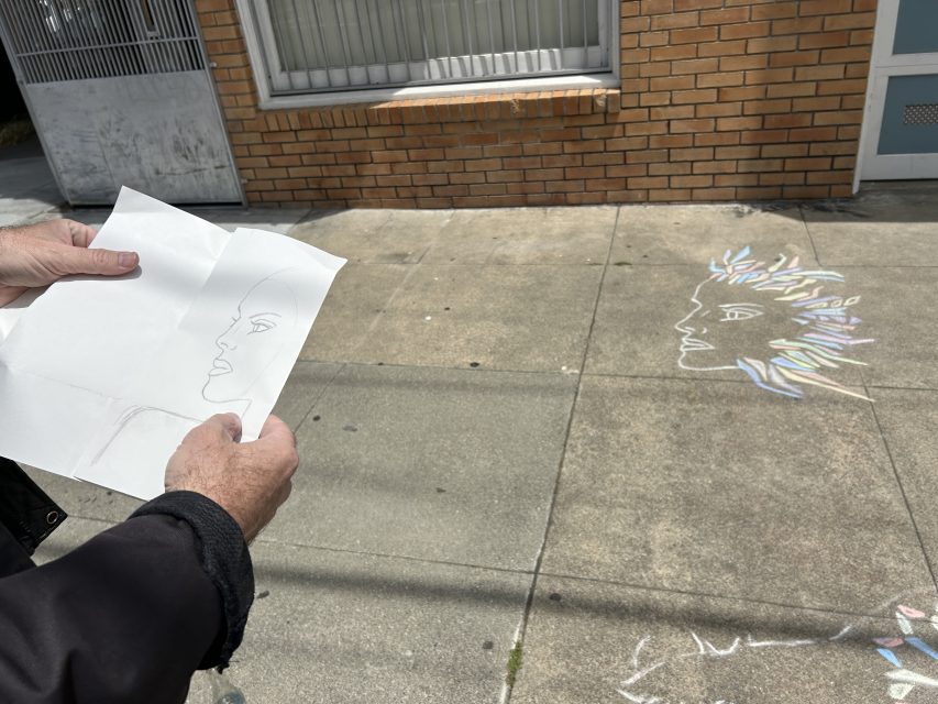 A person holds a pencil sketch of a face while looking at a colorful face illustration drawn on the sidewalk in front of a building.