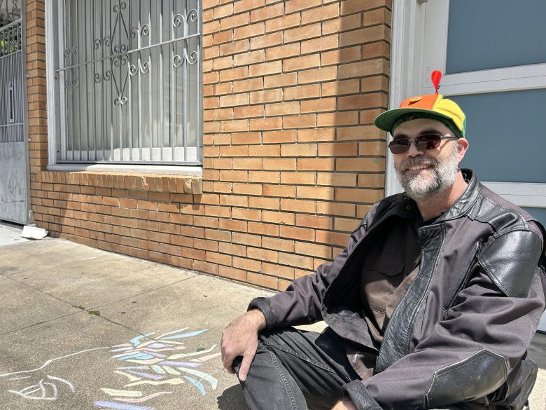A man wearing a propeller hat and sunglasses sits on a sidewalk next to chalk drawings, in front of a brick building with barred windows.