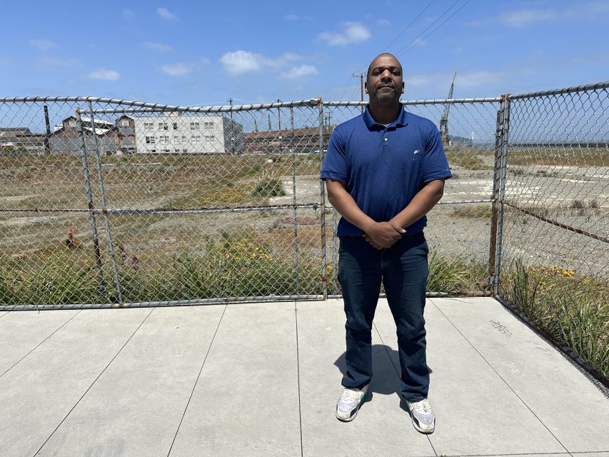 A man in a blue polo shirt and jeans stands on a sidewalk in front of a chain-link fence with industrial buildings and grass in the background under a partly cloudy sky.
