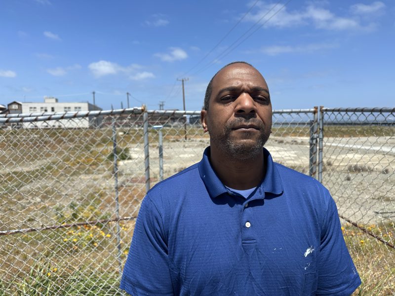 A man in a blue polo shirt stands in front of a chain-link fence with an industrial area and blue sky in the background.