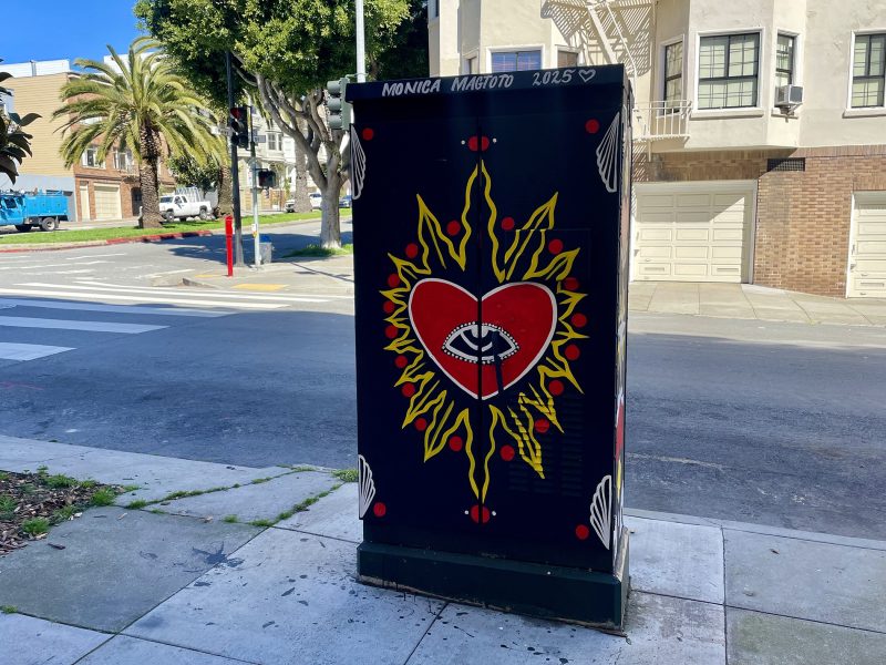 A utility box painted with a red heart, an eye in the center, surrounded by yellow rays and red teardrop shapes; "Monica Magoto 2025" is written at the top.
