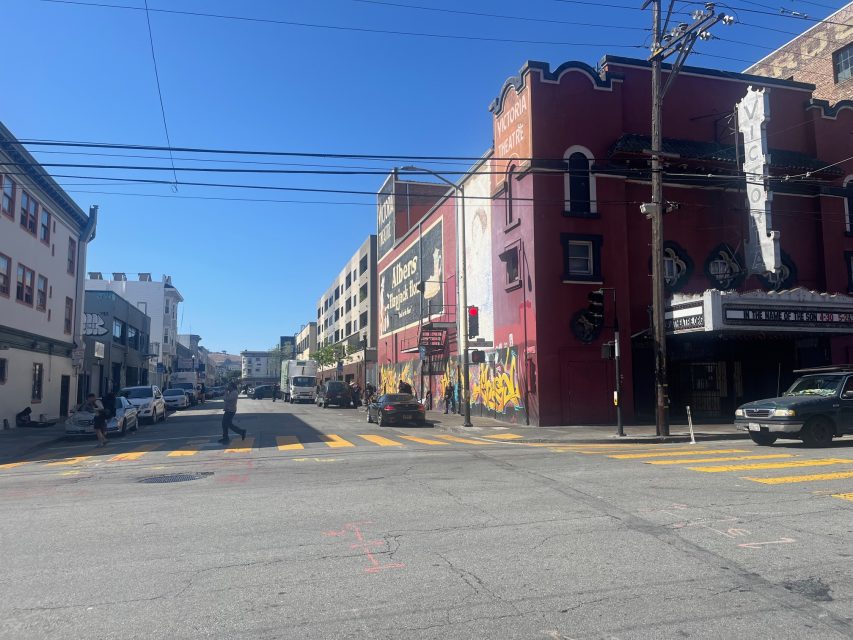A city street intersection at 16th and Mission with crosswalks, buildings, parked cars, and a red building featuring a mural and marquee sign under a clear blue sky.