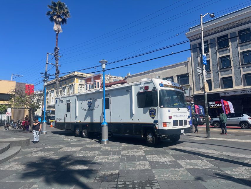 A large police mobile command vehicle is parked on a city plaza at 16th and Mission, surrounded by buildings under a clear blue sky.