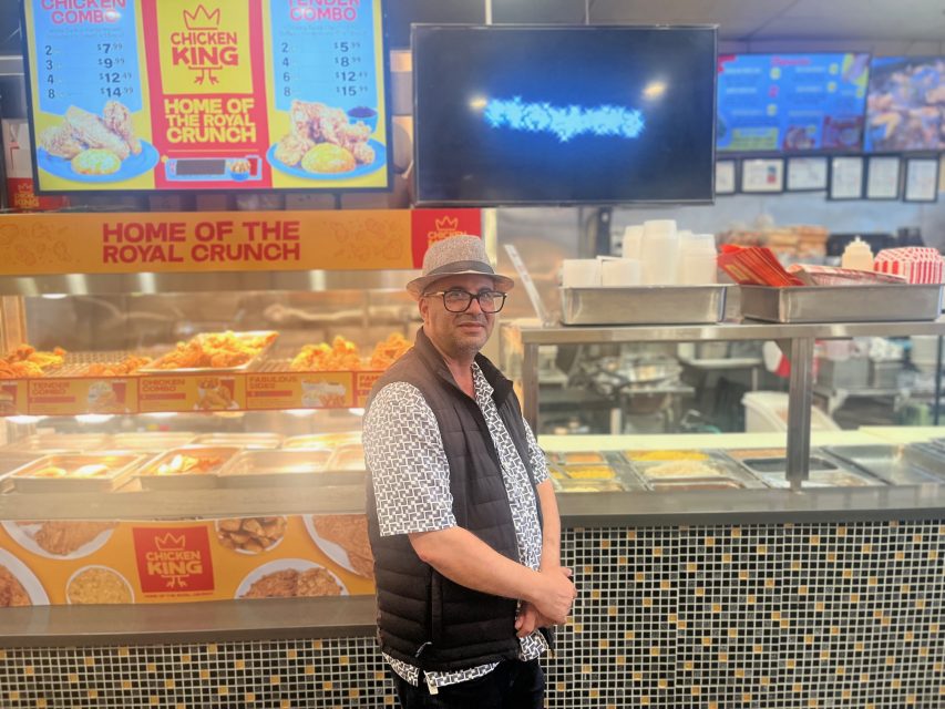 A man wearing glasses and a hat stands in front of a Chicken King counter at 16th and Mission, with menu boards and fried food visible in the background.