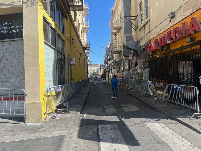 A narrow urban alley near 16th and Mission is blocked by metal barricades, with a person standing in the center. Buildings with graffiti and business signs line both sides under a clear sky.