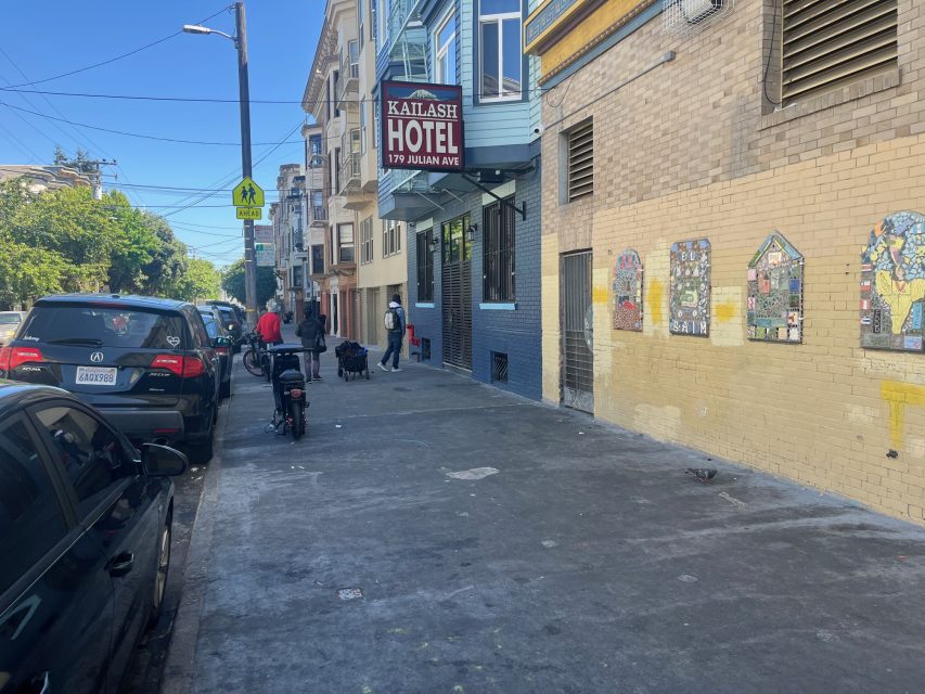 A sidewalk near 16th and Mission on Julian Ave with the Katlash Hotel sign above, people walking by parked cars, and mosaic art brightening a yellow wall.