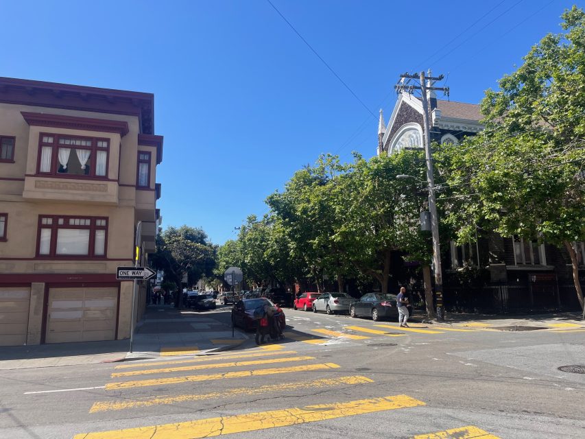 A sunny street intersection at 16th and Mission with yellow crosswalk lines, parked cars, a "One Way" sign, and buildings surrounded by leafy trees.